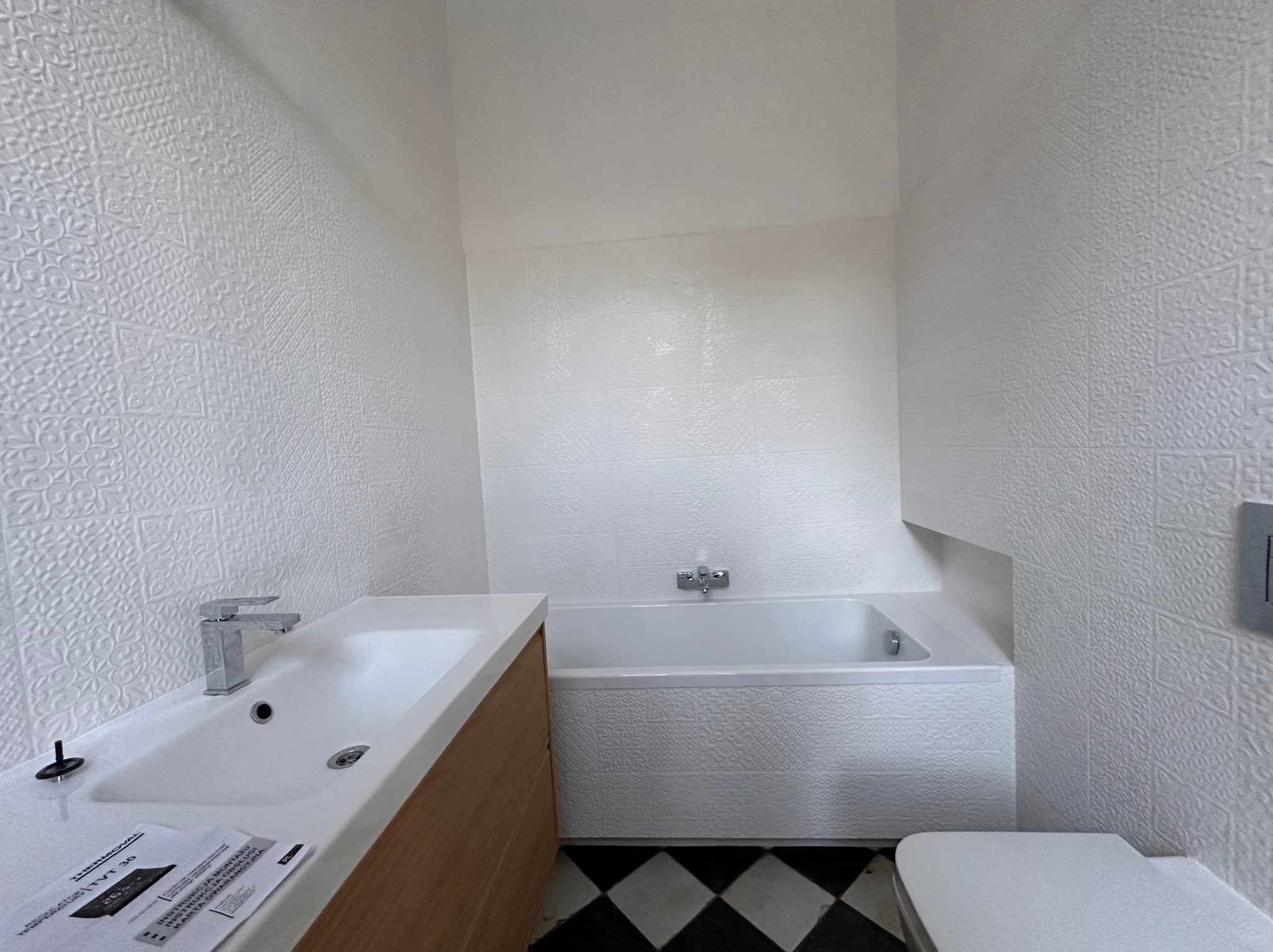 Nowoczesna łazienka z białą armaturą, wzorzystymi płytkami ściennymi i czarno-białą podłogą w szachownicę, widoczne instrukcje montażu na umywalce.