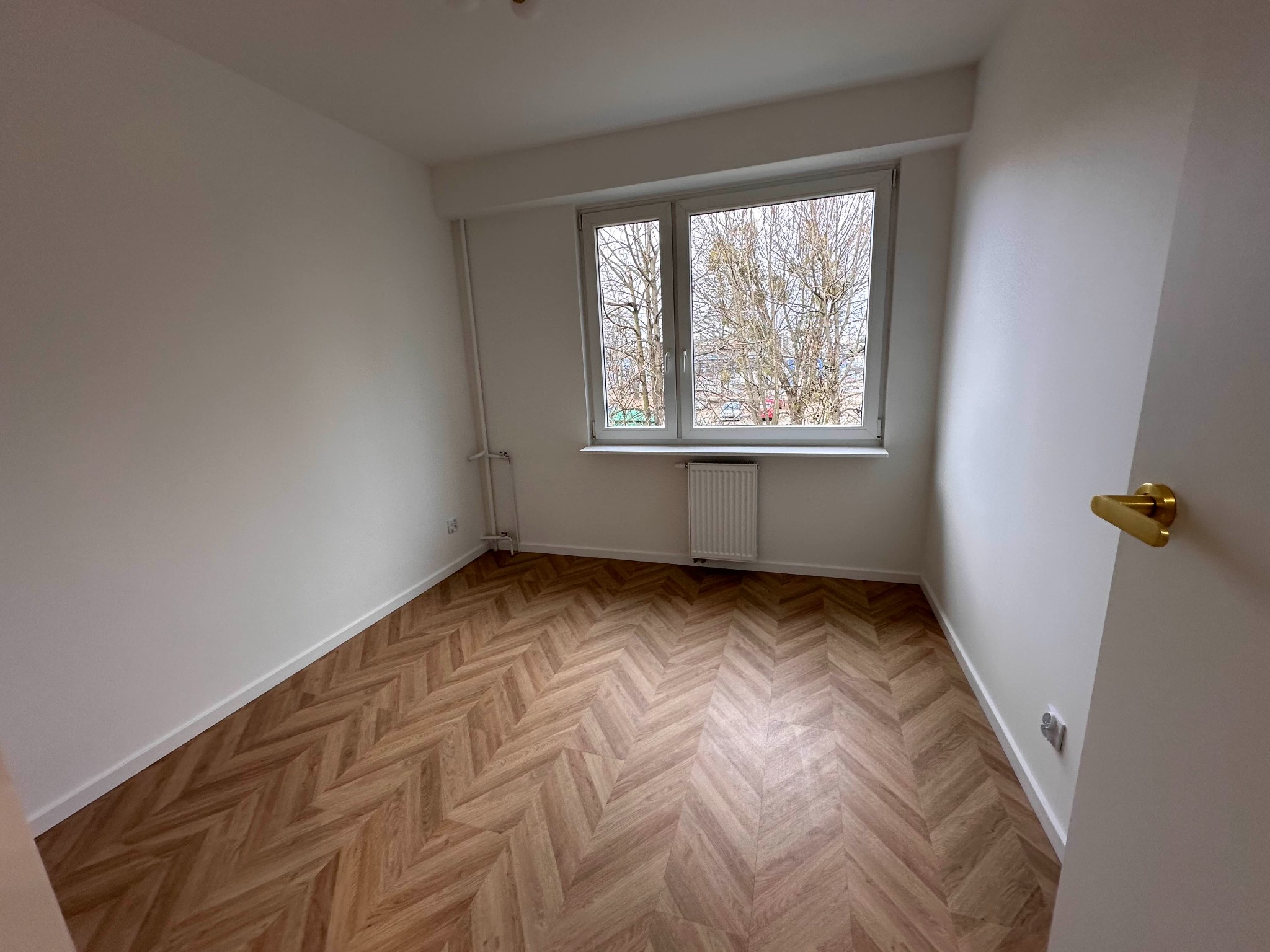 Pusty pokój po remoncie z parkietem ułożonym w jodełkę, białe ściany, okno z widokiem na drzewa bez liści, grzejnik pod oknem, złota klamka przy białych drzwiach.
