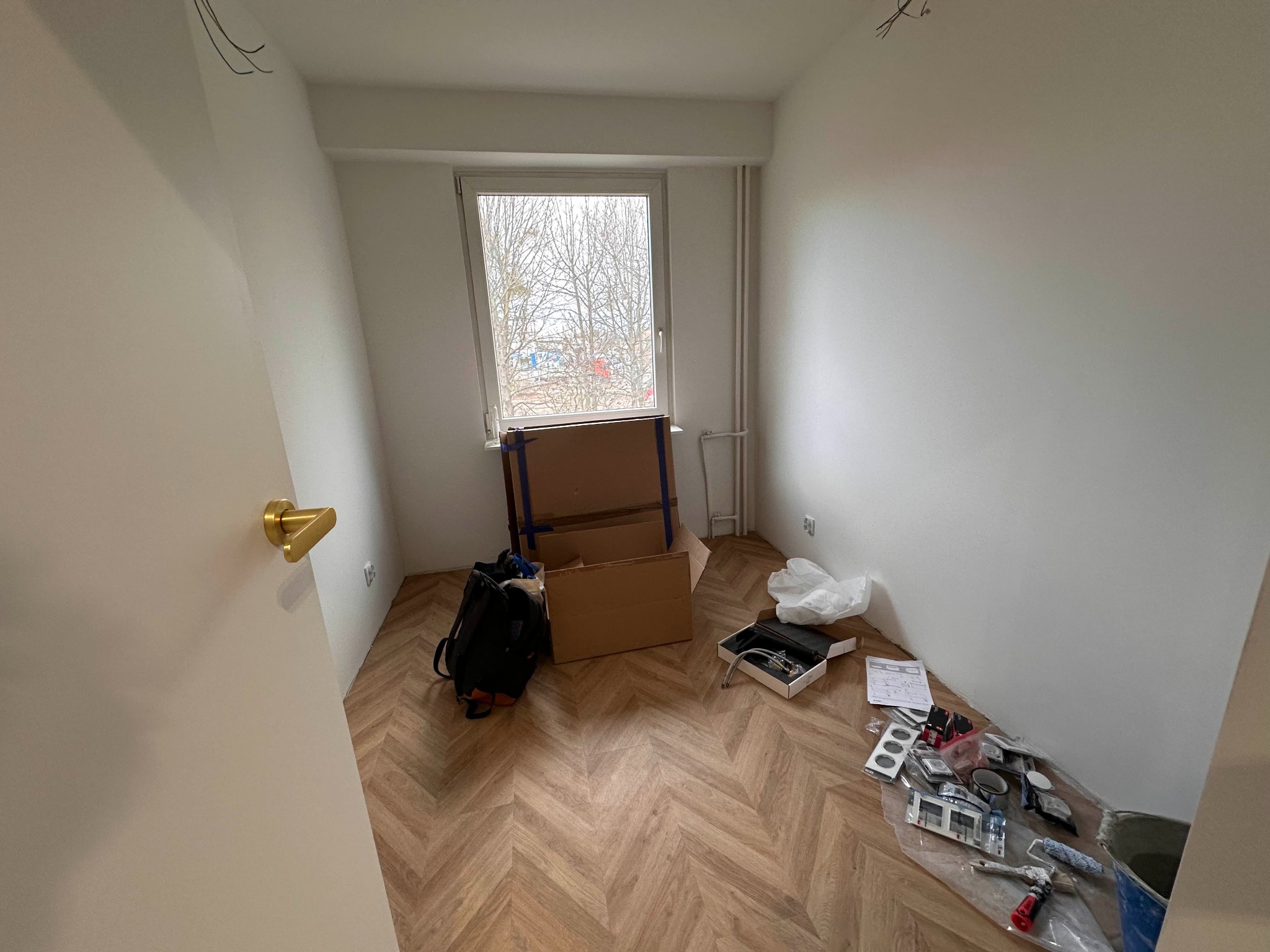 Pokój z nową podłogą w jodełkę, kartonami, plecakiem i narzędziami przygotowanymi do dalszych prac, widok na okno z drzewami bez liści.