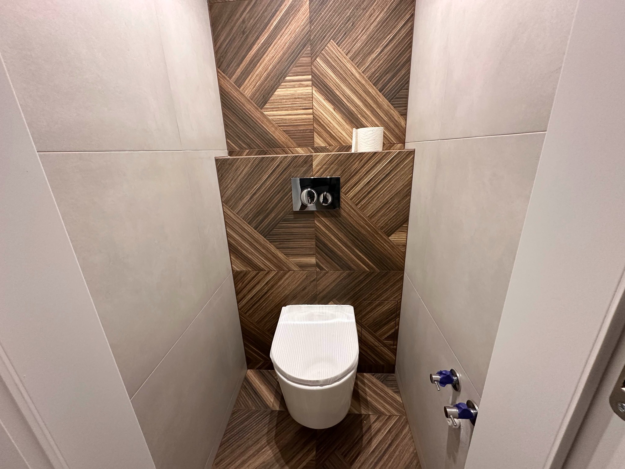 Nowoczesna toaleta z drewnopodobnymi płytkami ułożonymi w geometryczny wzór na ścianie i podłodze, biała ceramika sanitarna, ukryta spłuczka z chromowanymi przyciskami.
