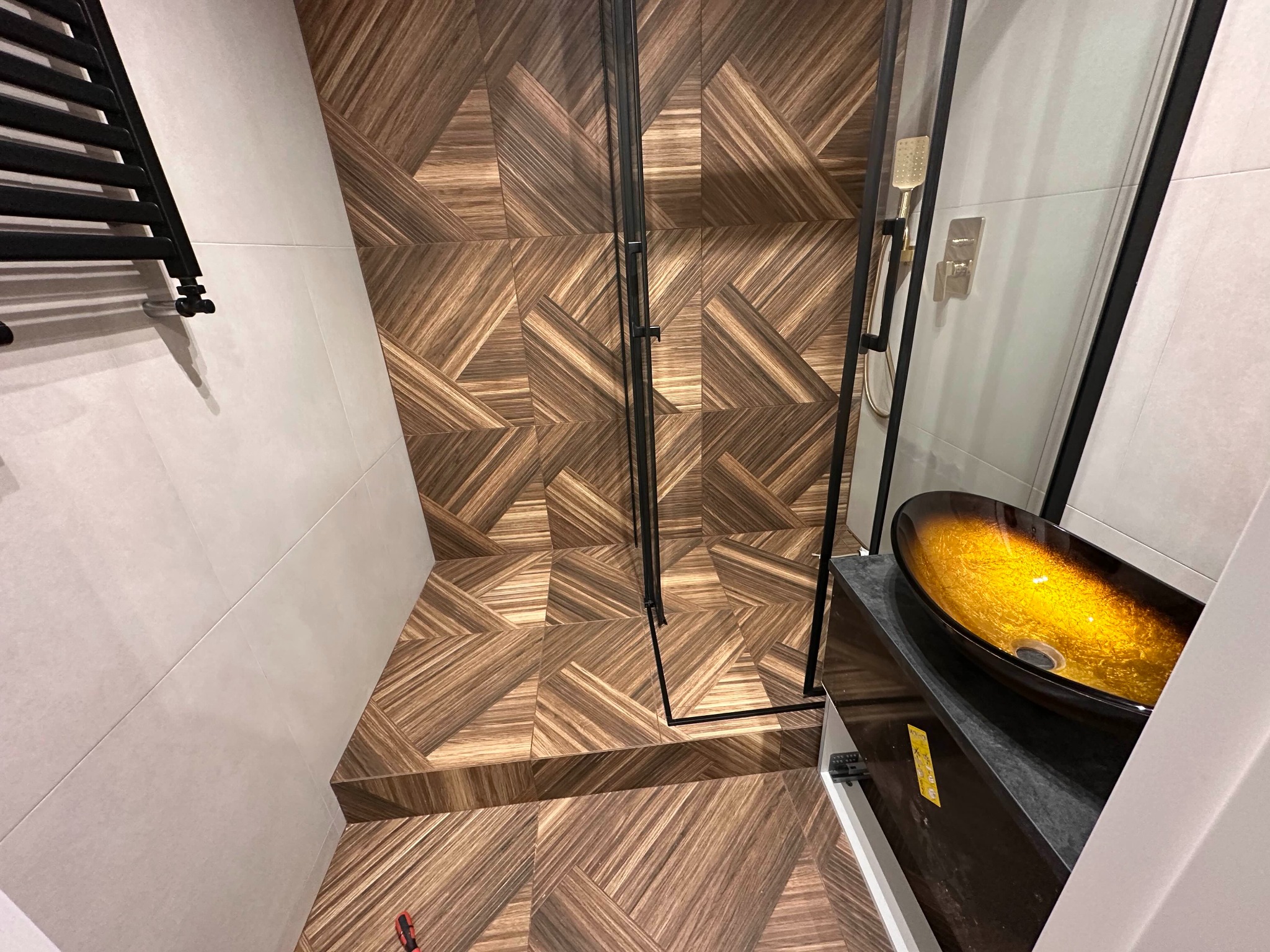 Nowoczesna łazienka z kabiną prysznicową typu walk-in, wykończona płytkami imitującymi drewno ułożonymi w geometryczny wzór, oraz umywalką nablatową w odcieniach bursztynu.