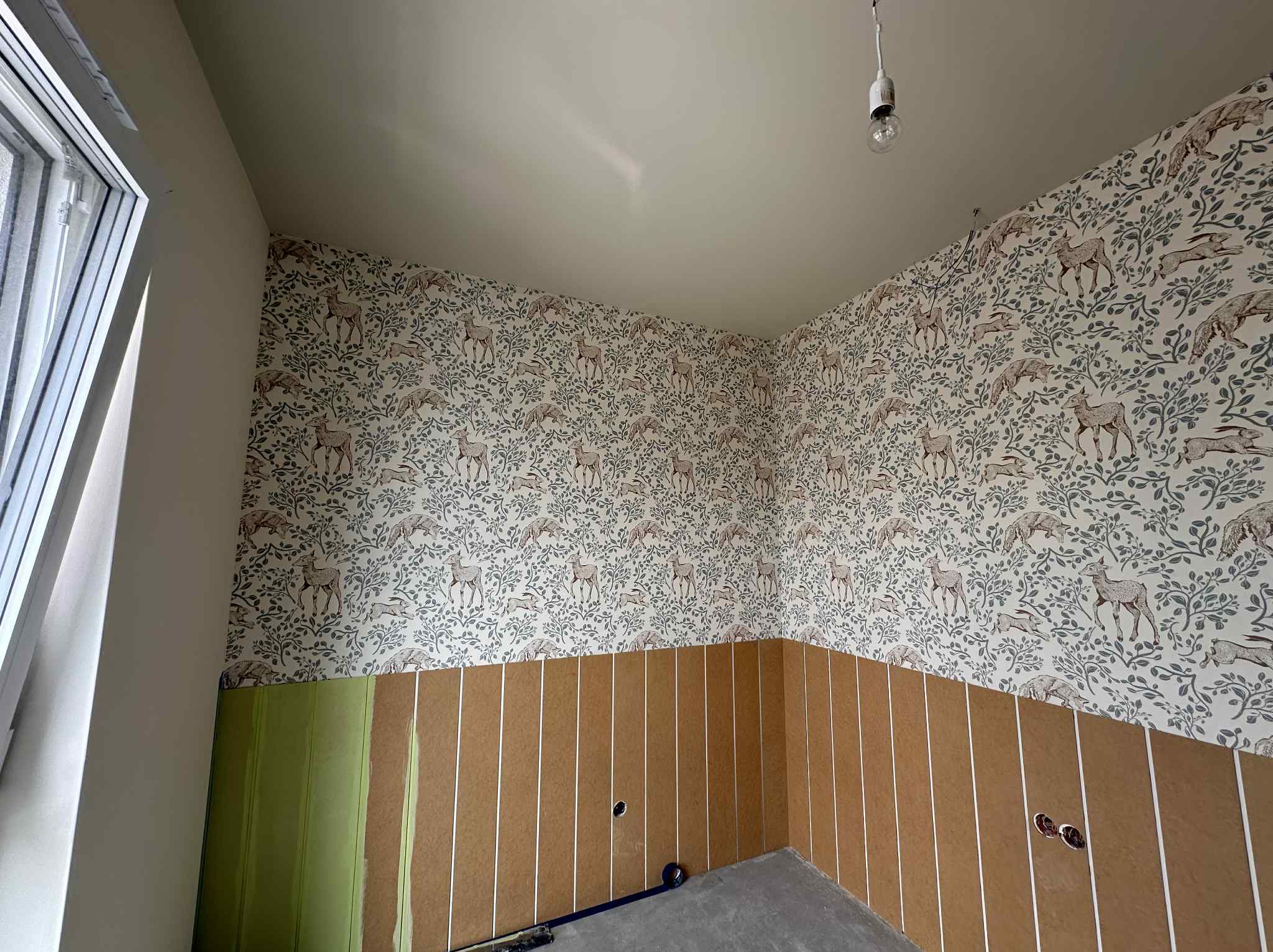 Pokój w trakcie wykańczania: tapeta w leśny motyw na górze, panele ścienne na dole. Widoczne gniazdka elektryczne i okno dachowe. Oświetlenie sufitowe tymczasowe.