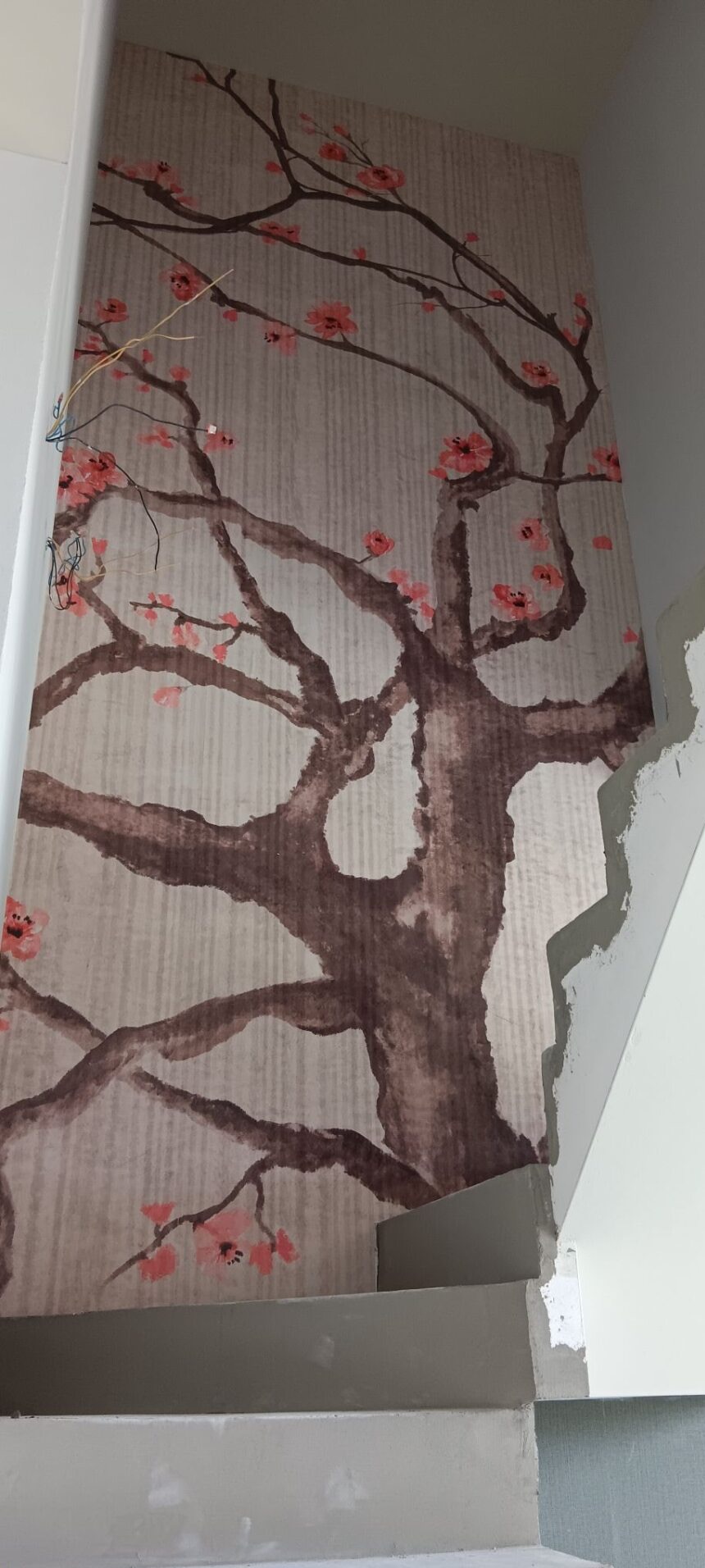 Tapeta z motywem kwitnącej wiśni na ścianie w trakcie wykańczania, widoczne przewody elektryczne i fragment surowej ściany. Dekoracyjne wykończenie wnętrza.