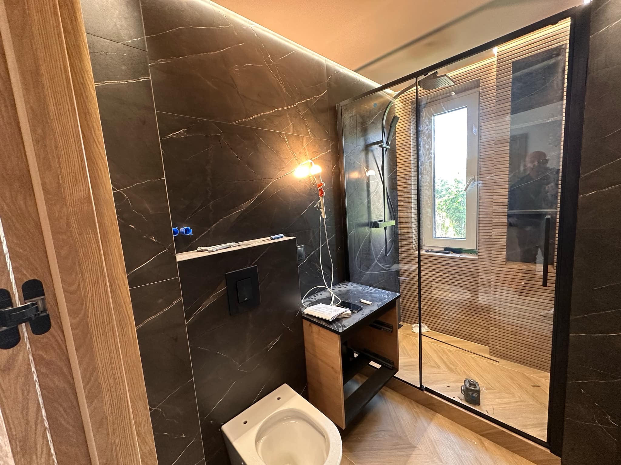 Nowoczesna łazienka w trakcie wykańczania: marmurowe ściany, drewniane akcenty, kabina prysznicowa z czarnymi ramami, wisząca toaleta i narzędzia pracy.