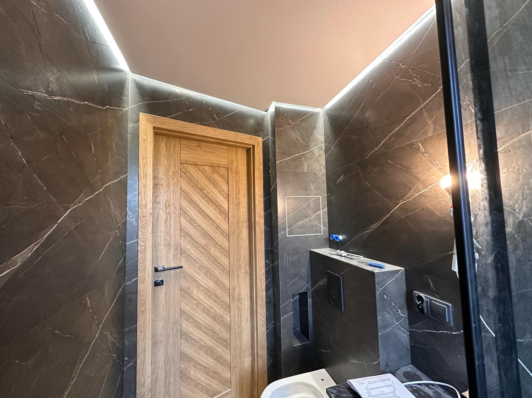 Wykończona łazienka z czarnymi marmurowymi płytkami, drewnianymi drzwiami z wzorem w jodełkę i podświetleniem LED. Widoczne elementy instalacji sanitarnej.
