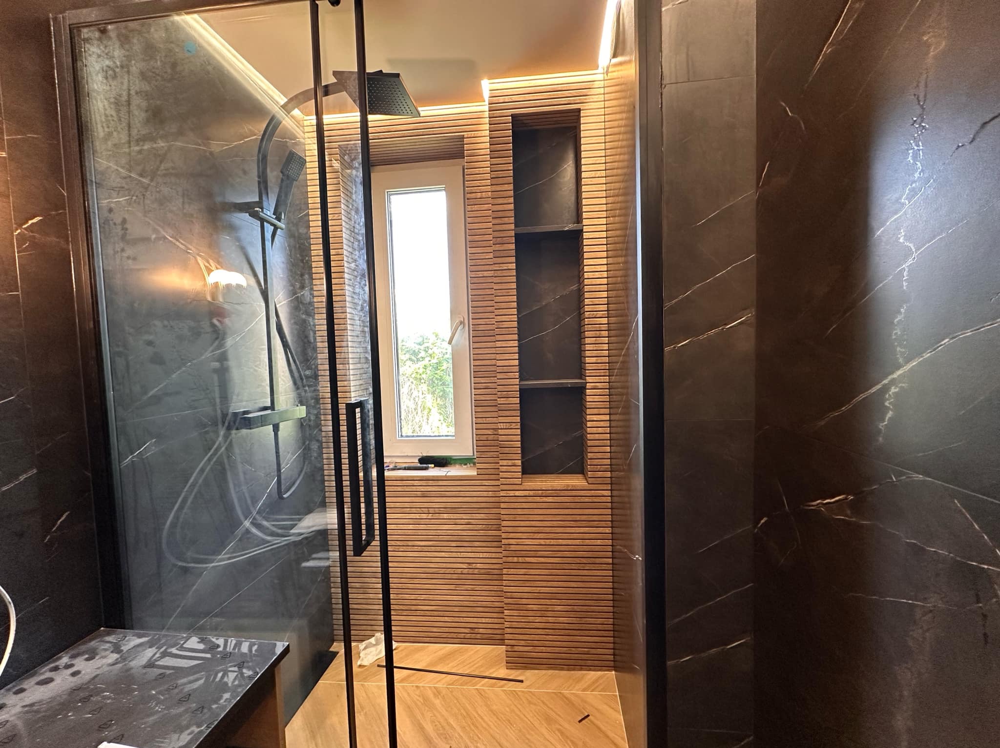 Nowoczesna kabina prysznicowa z czarnymi płytkami imitującymi marmur i drewnianą okładziną. W tle okno i wnęka z półkami. Oświetlenie LED podkreśla detale.