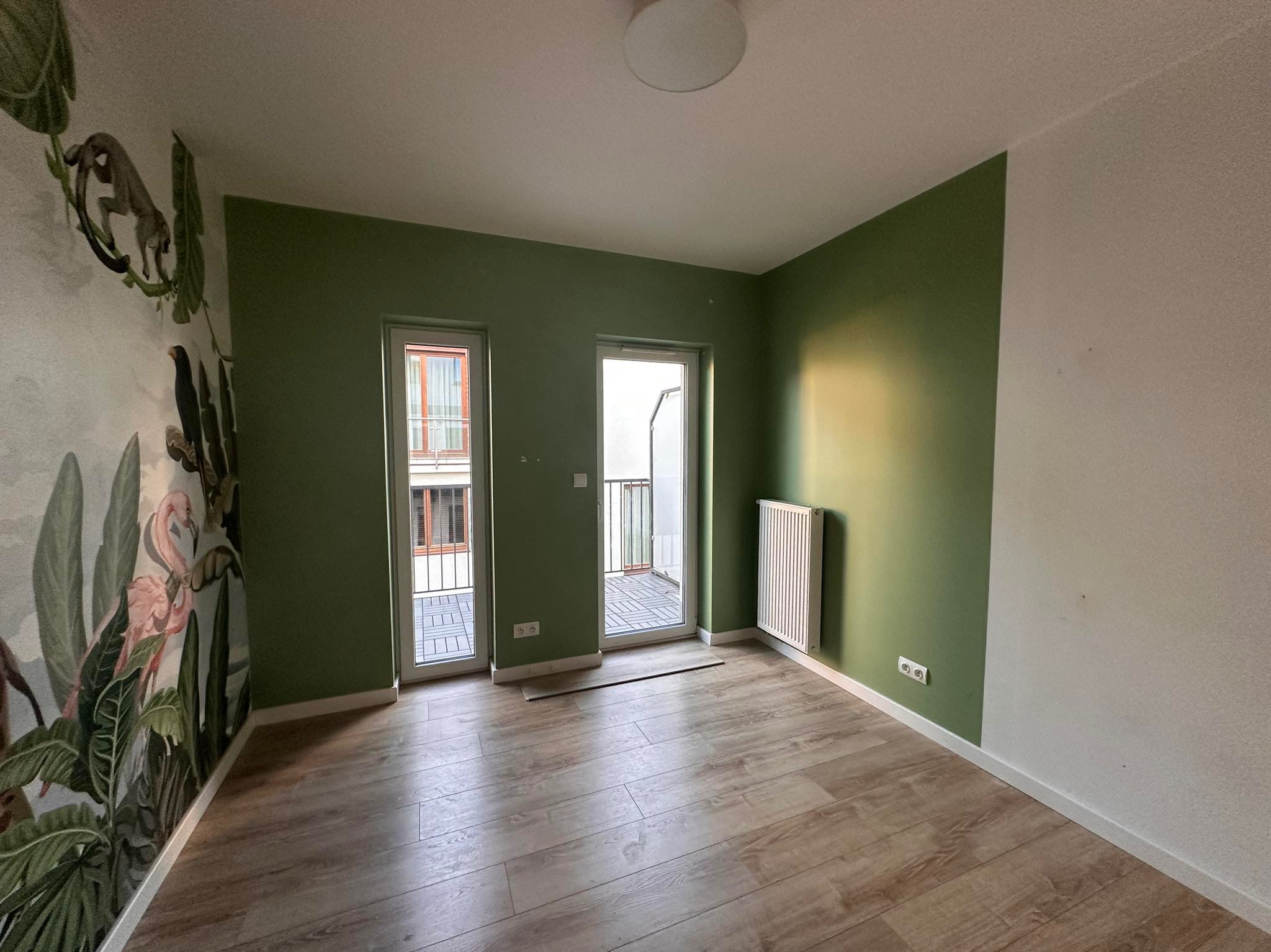 Pokój po malowaniu z zielonymi ścianami, drewnianą podłogą i fototapetą z motywem dżungli. Widoczne okna balkonowe i grzejnik. Świeżo wykończone wnętrze.