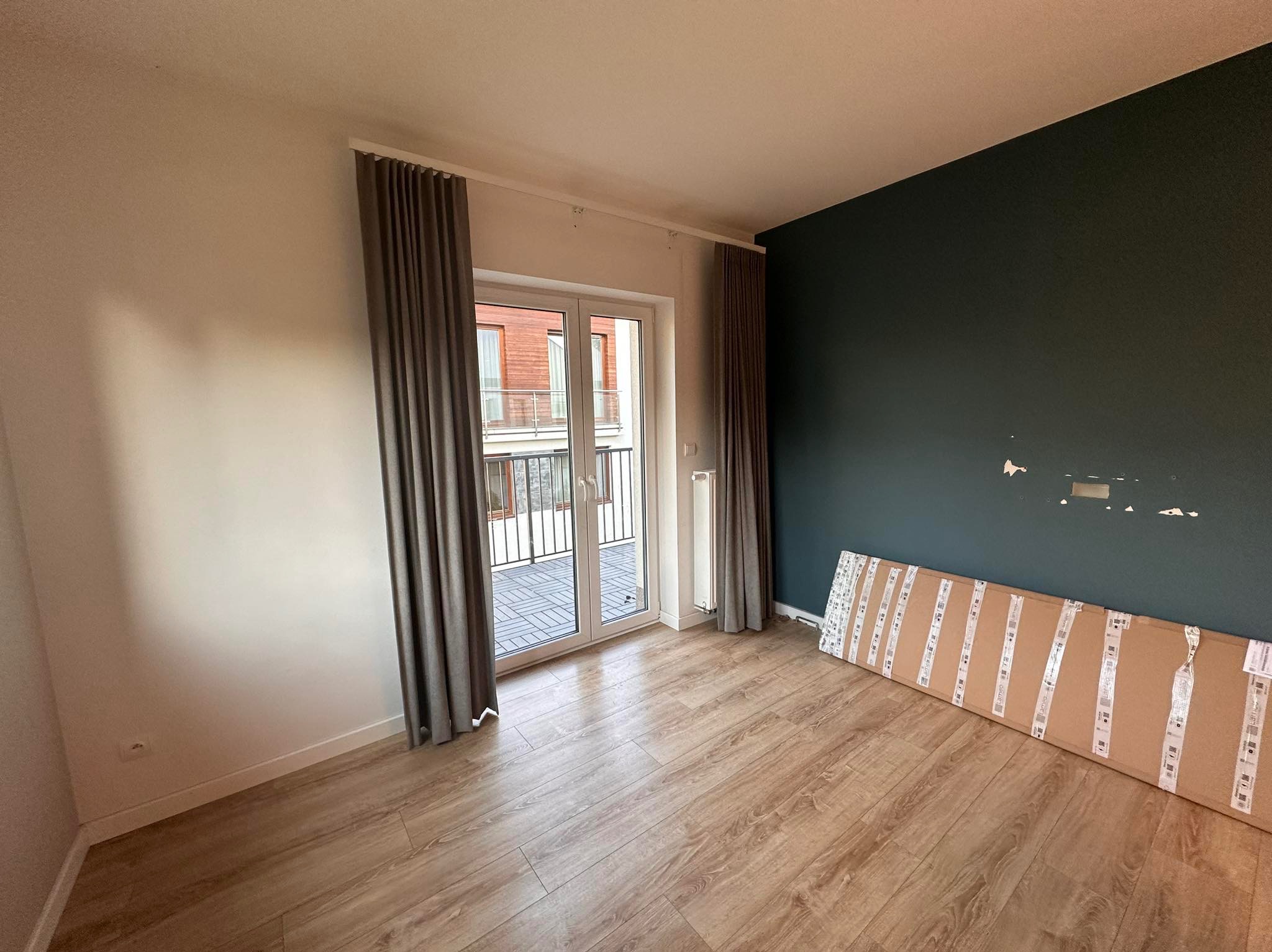 Pokój po remoncie: drewniana podłoga, ciemna ściana, okno balkonowe z szarymi zasłonami. Na podłodze leżą panele podłogowe w kartonach, gotowe do montażu.