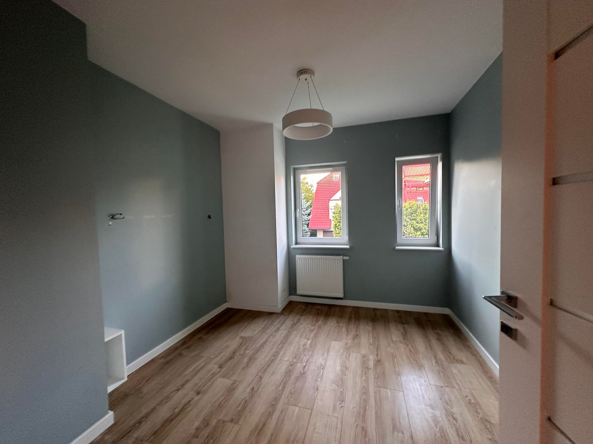 Pomalowane na szaro ściany, drewnopodobna podłoga i białe okna z grzejnikiem w pustym pokoju. Nowoczesna lampa sufitowa. Widok zza otwartych drzwi.