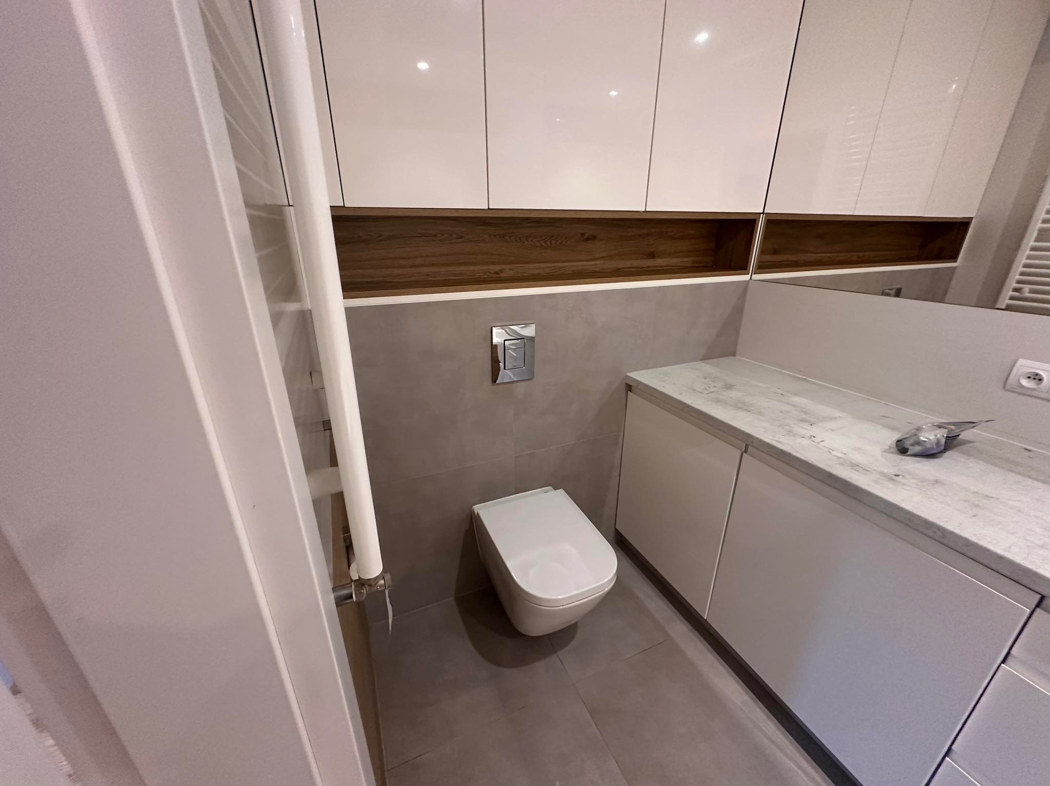 Nowoczesna toaleta w szaro-białej kolorystyce z podwieszaną miską WC, zabudową meblową i grzejnikiem drabinkowym. Widoczne płytki ceramiczne i drewniane akcenty.