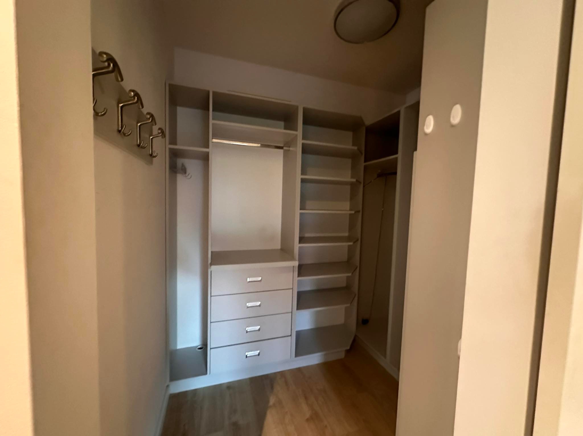 Wnętrze garderoby z szarymi szafkami, półkami i czterema szufladami. Na ścianie wieszaki w kształcie ludzików. Widoczny fragment podłogi.