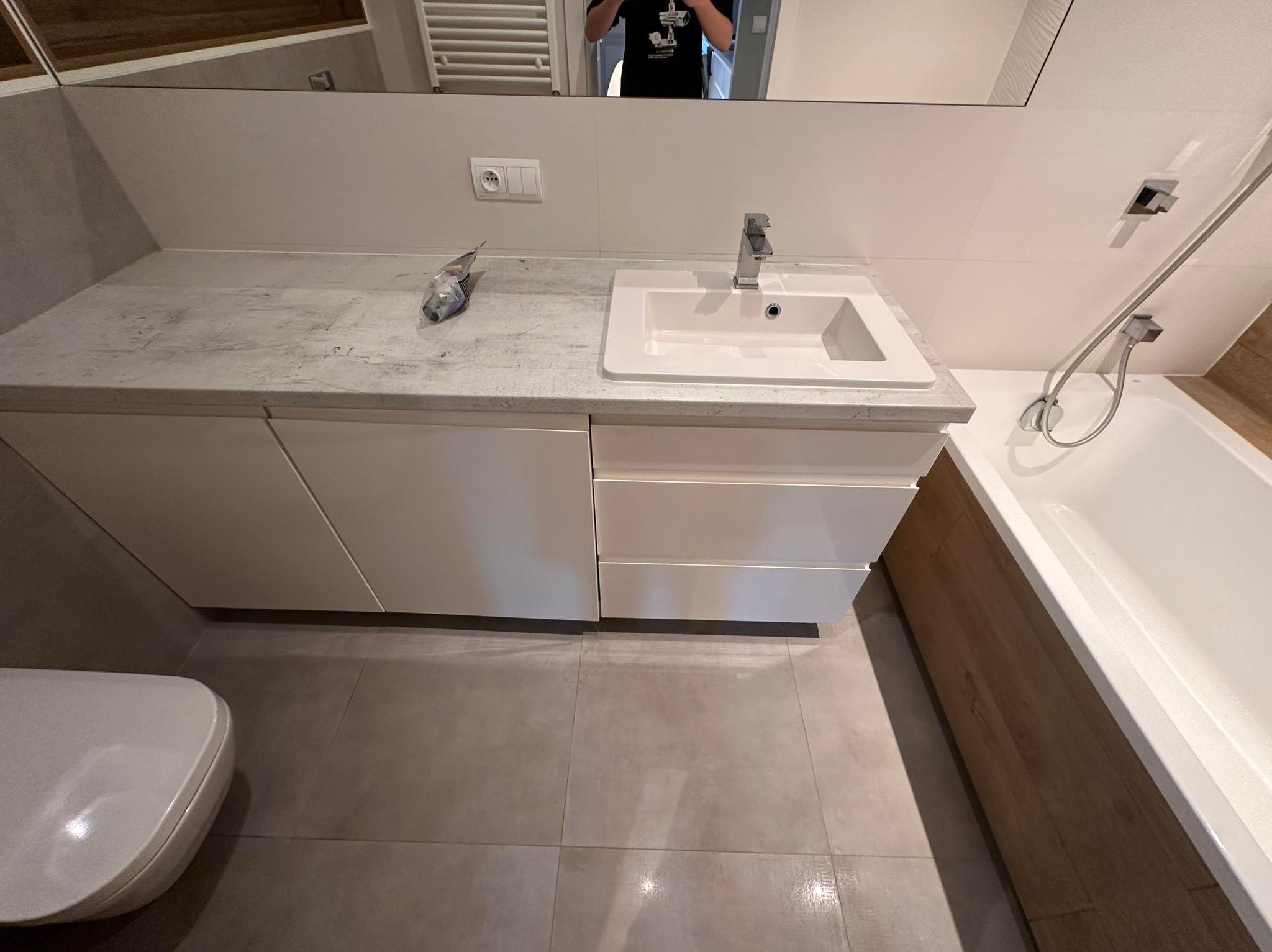 Nowoczesna łazienka po remoncie: białe szafki pod umywalką z szarym blatem, bateria, toaleta i wanna z drewnianą obudową. Widoczne płytki podłogowe.