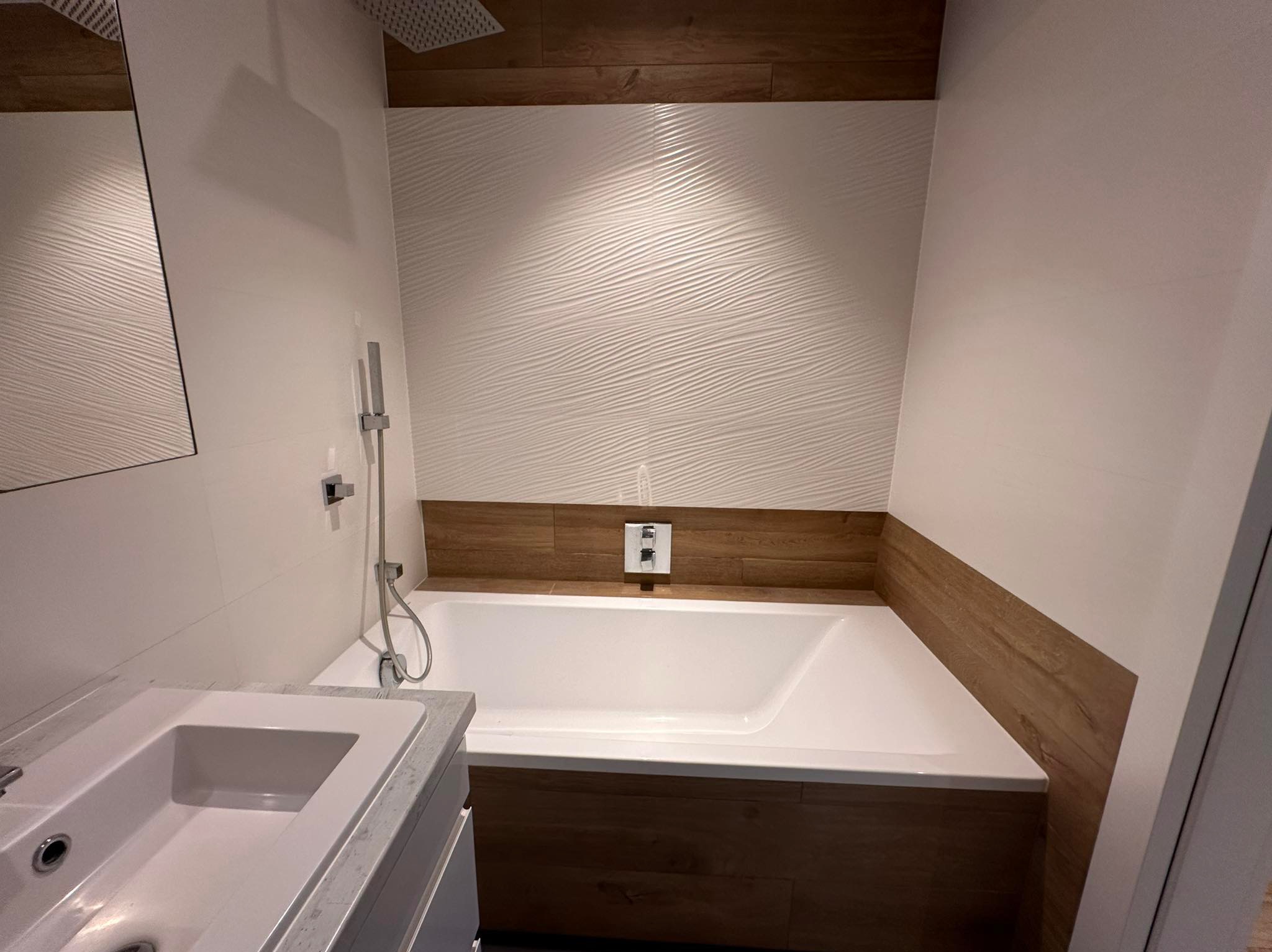 Nowoczesna łazienka po metamorfozie: biała wanna obudowana drewnem, umywalka z szarą armaturą, dekoracyjne płytki 3D na ścianie i deszczownica.