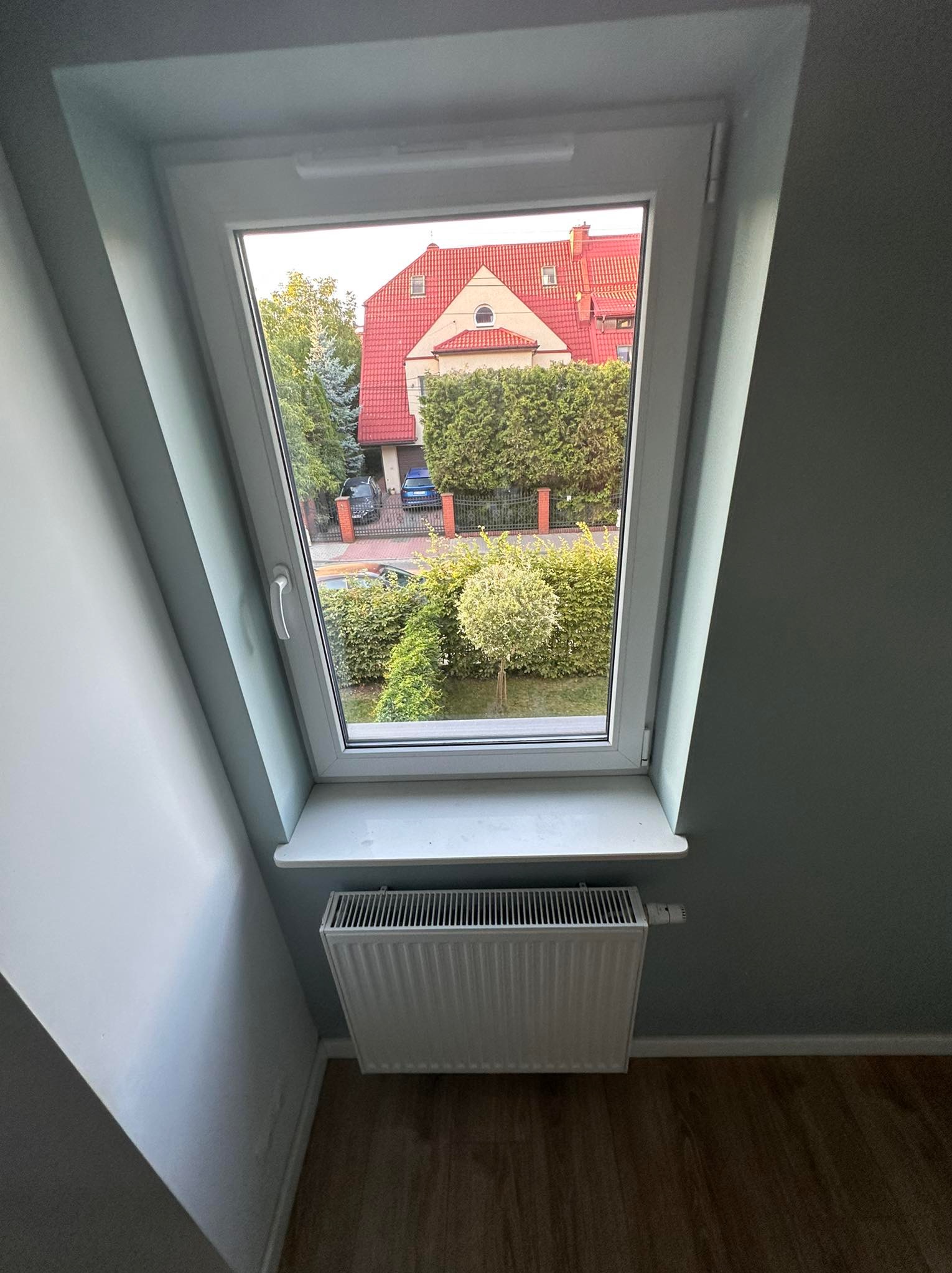 Białe okno dachowe z widokiem na dom z czerwoną dachówką i zadbany ogród. Pod oknem biały grzejnik. Ściany w odcieniach szarości. Ujęcie z dołu.