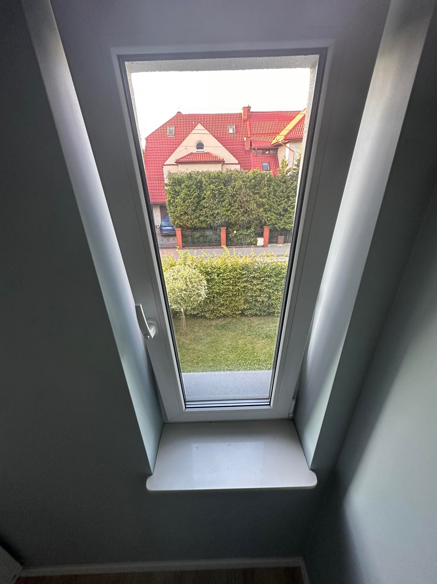 Białe okno dachowe z widokiem na dom z czerwoną dachówką i zielony ogród. Szare ściany i parapet wewnątrz. Ujęcie z dołu, podkreślające kąt nachylenia okna.