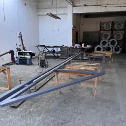 Warsztatowa sceneria z ramą bramy wjazdowej na drewnianych kozłach, spawarką i zwojami drutu w tle.