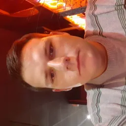 Portret mężczyzny w swetrze w paski, z widocznym oświetleniem sufitowym w tle. Ujęcie z bliska, z perspektywy żabiej.