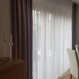 Eleganckie, brązowe i białe zasłony z delikatnej tkaniny, upięte na karniszu, w tle widoczne okno i fragment jadalni z drewnianą podłogą i krzesłami.