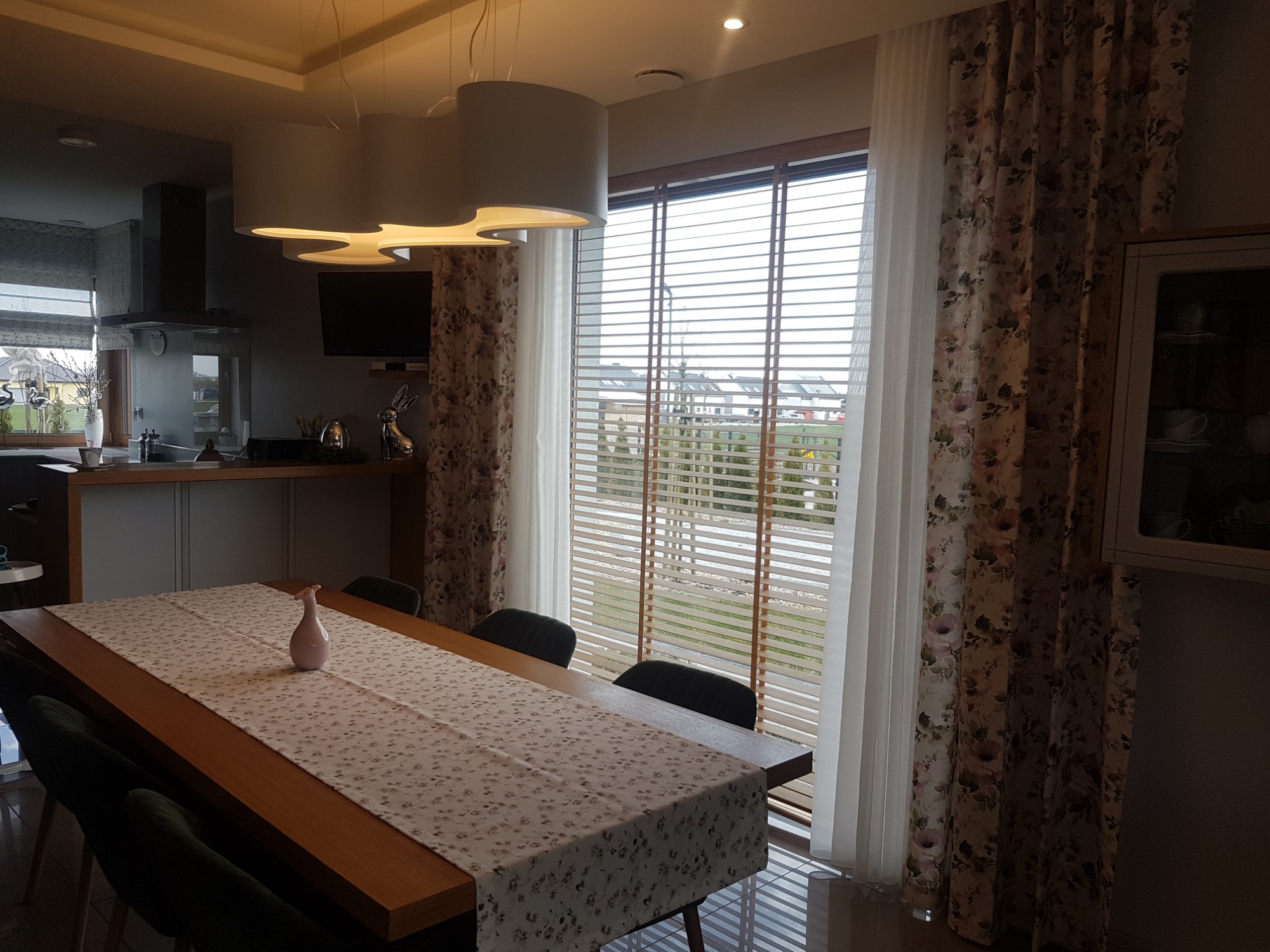Jadalnia w Jasin z drewnianym stołem i krzesłami, okno z drewnianymi żaluzjami poziomymi i zasłonami w kwiaty, nowoczesne lampy wiszące.