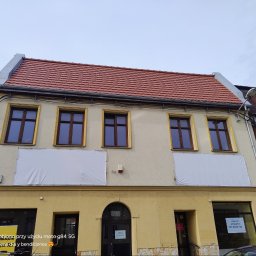 Red - Odnowiony budynek z czerwoną dachówką, widoczne rynny i okna z brązowymi ramami. Na elewacji banery reklamowe. Ujęcie z dołu, pochmurne niebo.