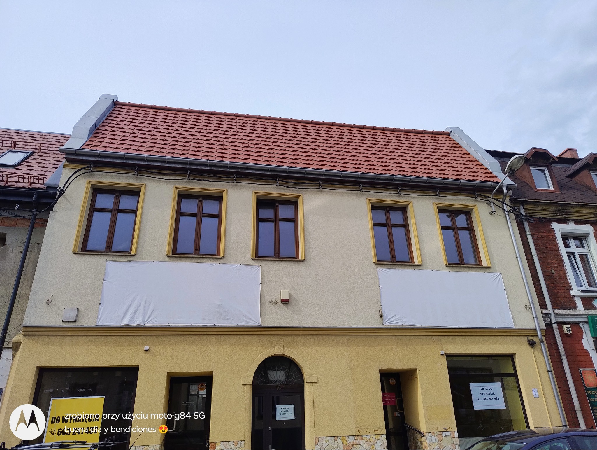 Odnowiony budynek z czerwoną dachówką, widoczne rynny i okna z brązowymi ramami. Na elewacji banery reklamowe. Ujęcie z dołu, pochmurne niebo.