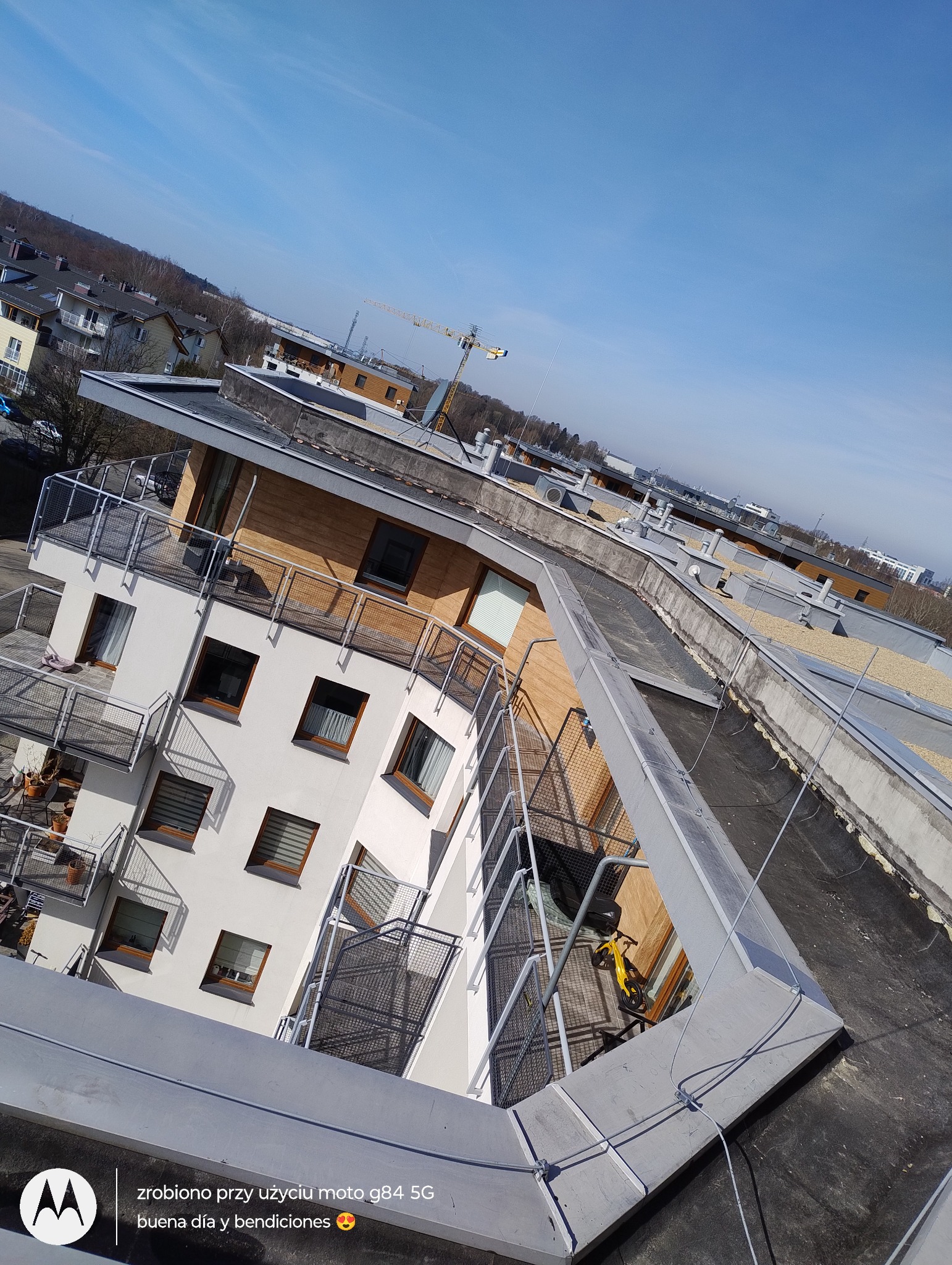 Widok z dachu na nowoczesny budynek z balkonami i elementami drewnianymi, w tle żuraw budowlany i błękitne niebo. Ujęcie z góry, podkreślające geometrię dachu.