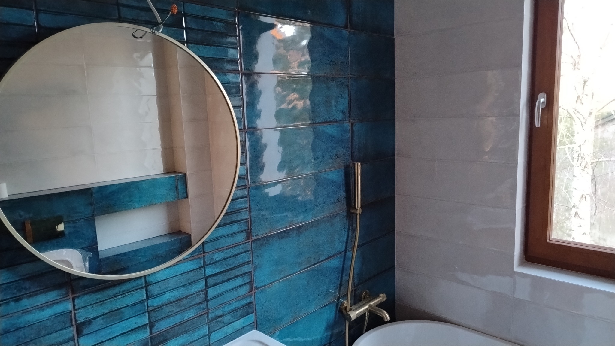 Nowoczesna łazienka z okrągłym lustrem, turkusowymi płytkami ułożonymi w cegiełkę i złotą armaturą. Widoczne białe płytki i drewniana rama okna.