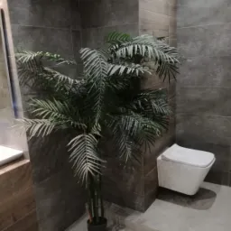 Nowoczesna łazienka z szarymi płytkami, wiszącą toaletą i drewnopodobną szafką pod umywalką, ożywiona sztuczną palmą w rogu