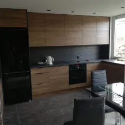 Nowoczesna kuchnia z drewnianymi frontami i czarnym blatem w Mysłowicach, widok na zabudowę kuchenną, lodówkę i stół z krzesłami.