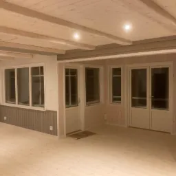 Jasne, drewniane wnętrze domu z oknami i drzwiami, wykończone w naturalnym stylu, widoczne listwy i belki stropowe, oświetlenie punktowe.