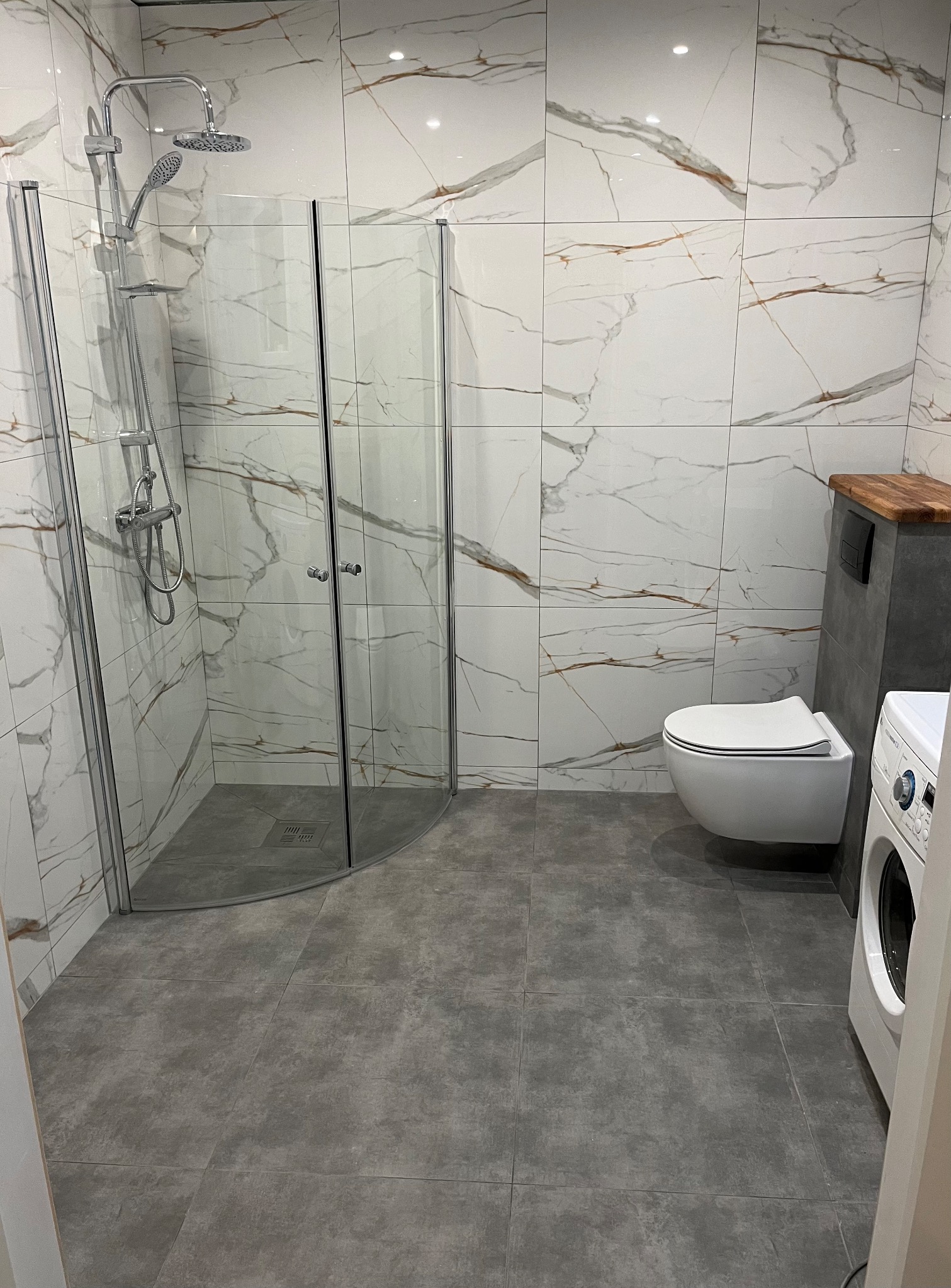 Nowoczesna łazienka z kabiną prysznicową typu walk-in, białą ceramiką i płytkami imitującymi marmur z brązowymi żyłami na ścianach, szara podłoga z dużych płytek.