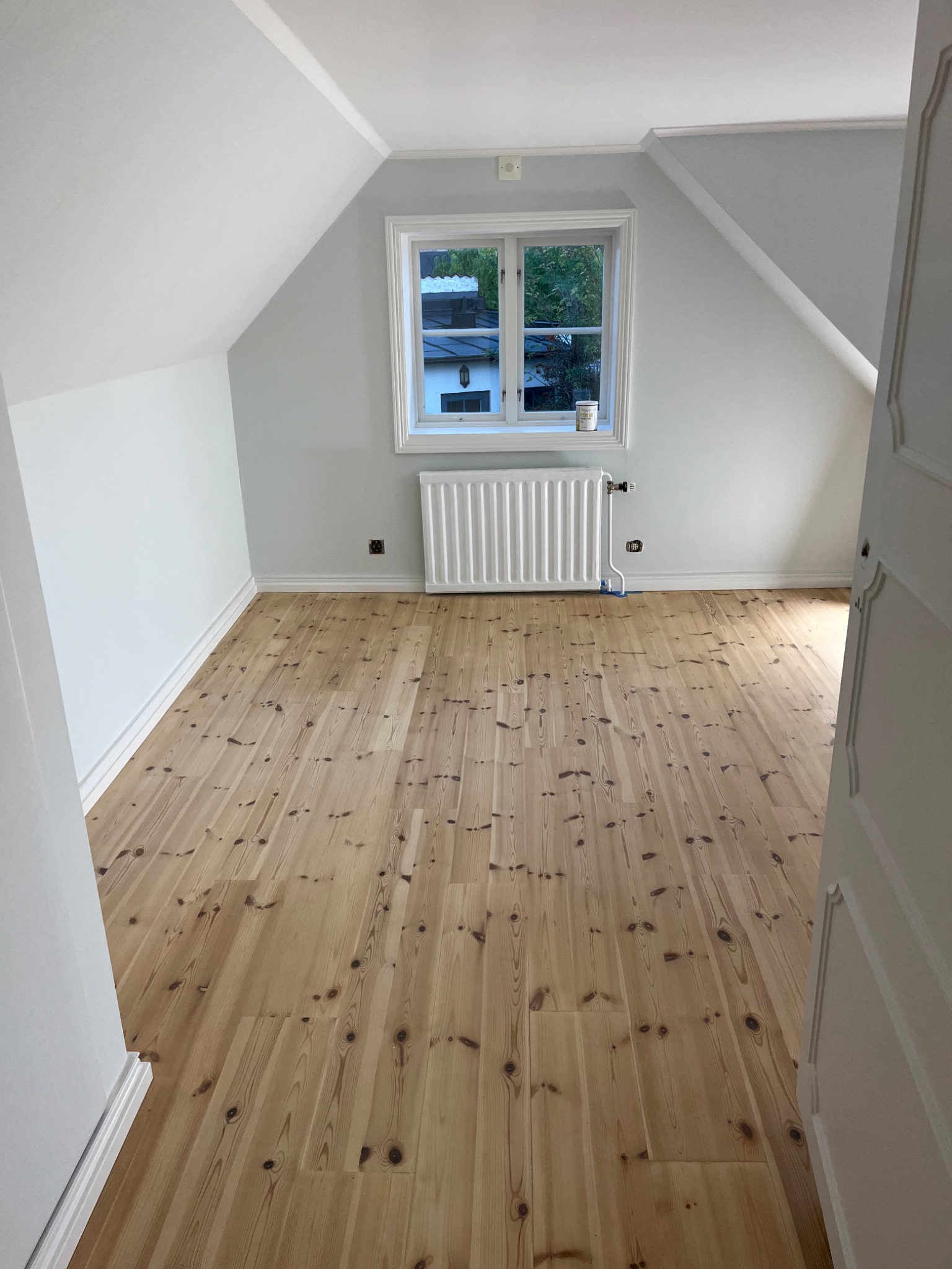 Poddasze po odświeżeniu: jasne ściany, drewniana podłoga z widocznymi sękami, biały grzejnik pod oknem z widokiem na zieleń.