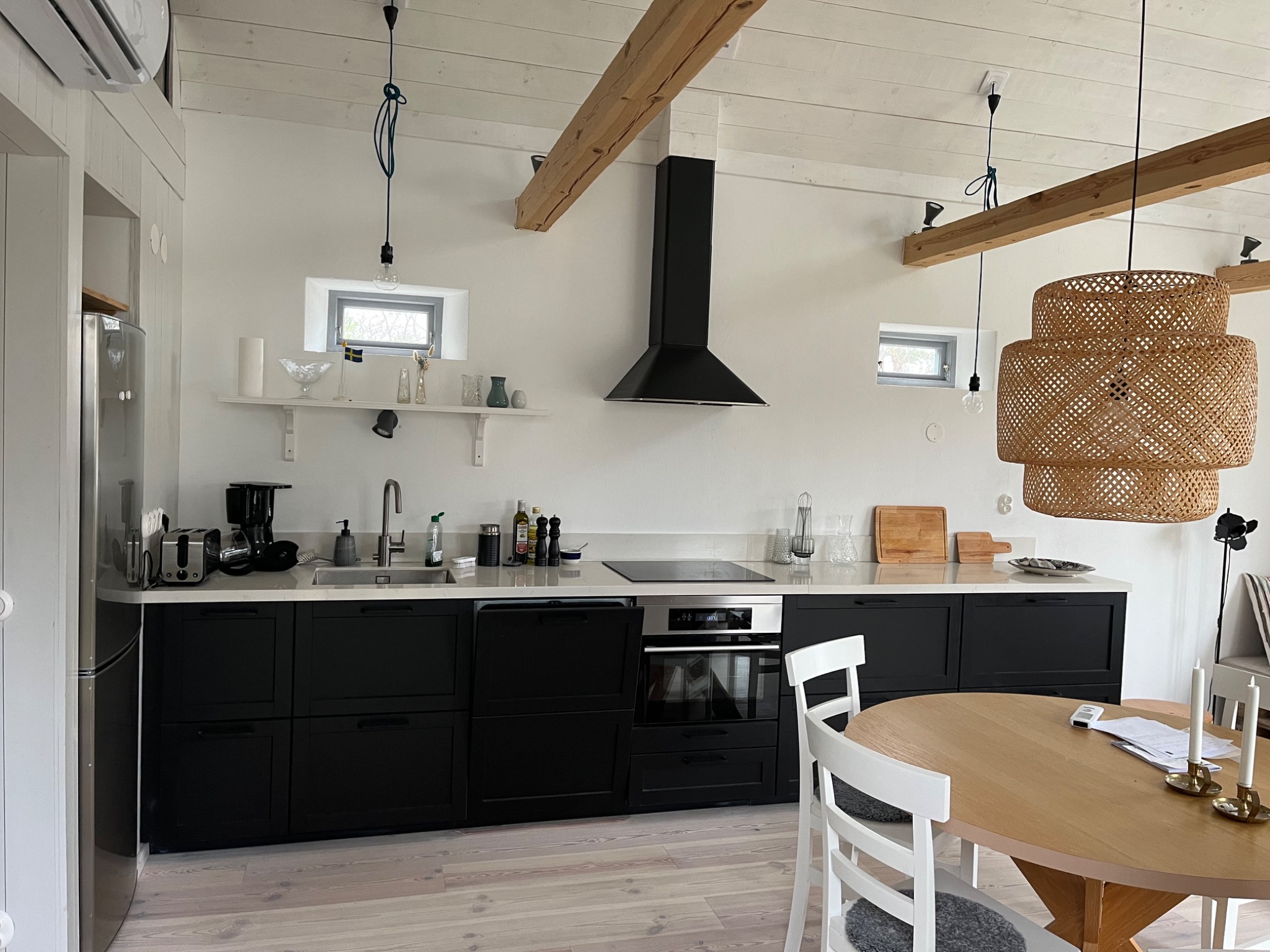 Jasna kuchnia w stylu skandynawskim z czarnymi szafkami, drewnianymi belkami stropowymi i okrągłym stołem z białymi krzesłami. Widoczny otwarty zmywarka i wisząca lampa z wikliny.