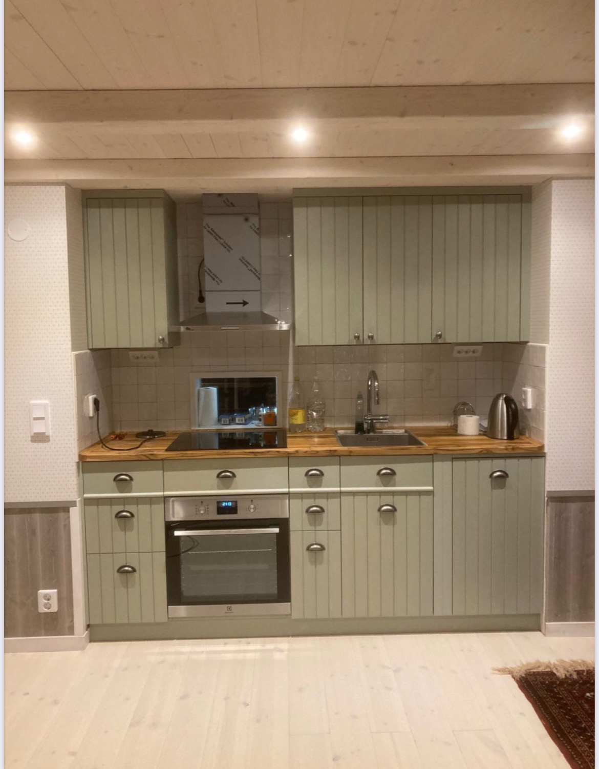 Jasna kuchnia z drewnianym blatem i frontami szafek w pastelowym odcieniu zieleni, z piekarnikiem elektrycznym i metalowym okapem.