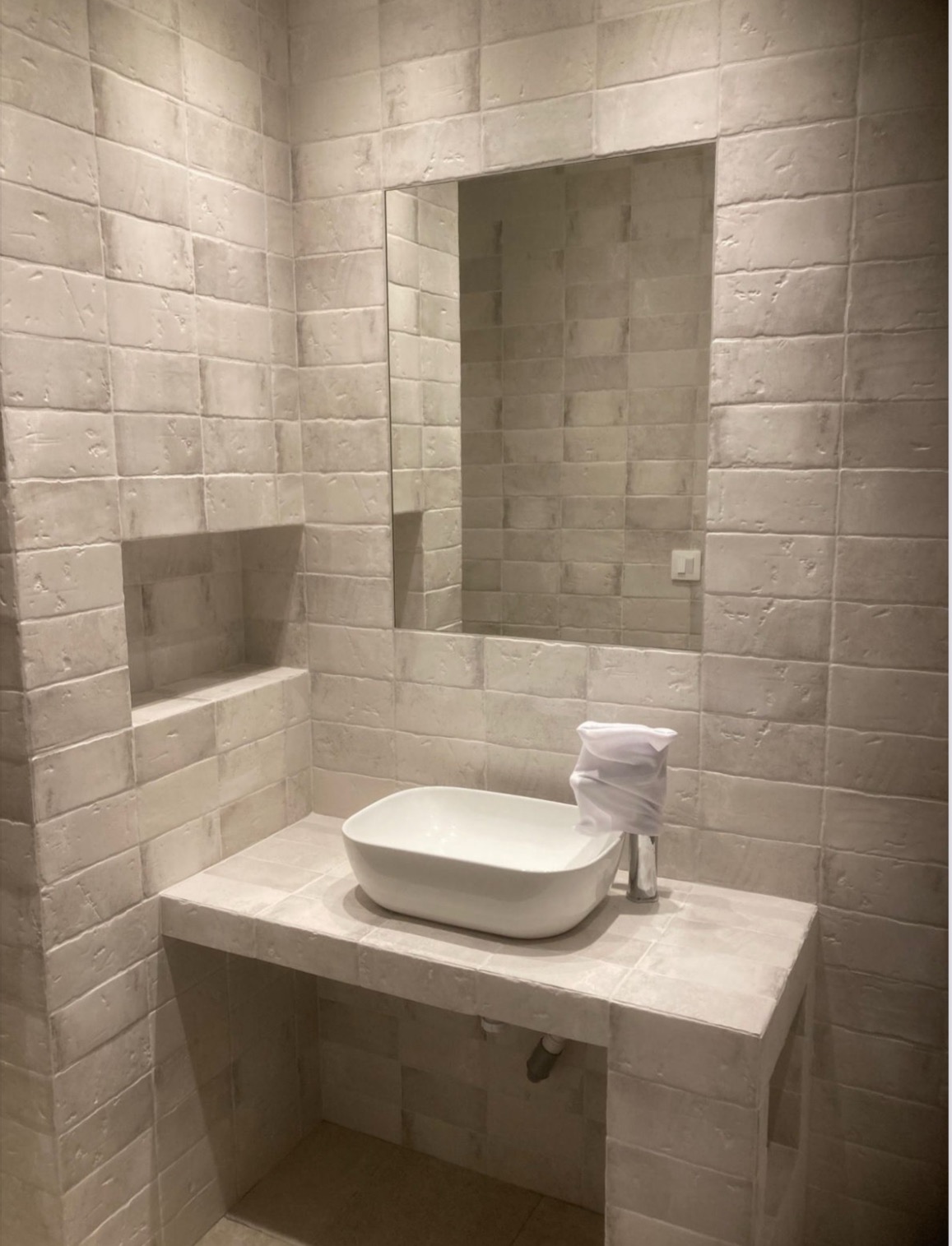 Nowoczesna łazienka z umywalką nablatową, lustrem i ścianami wyłożonymi beżowymi, strukturalnymi płytkami imitującymi cegłę.