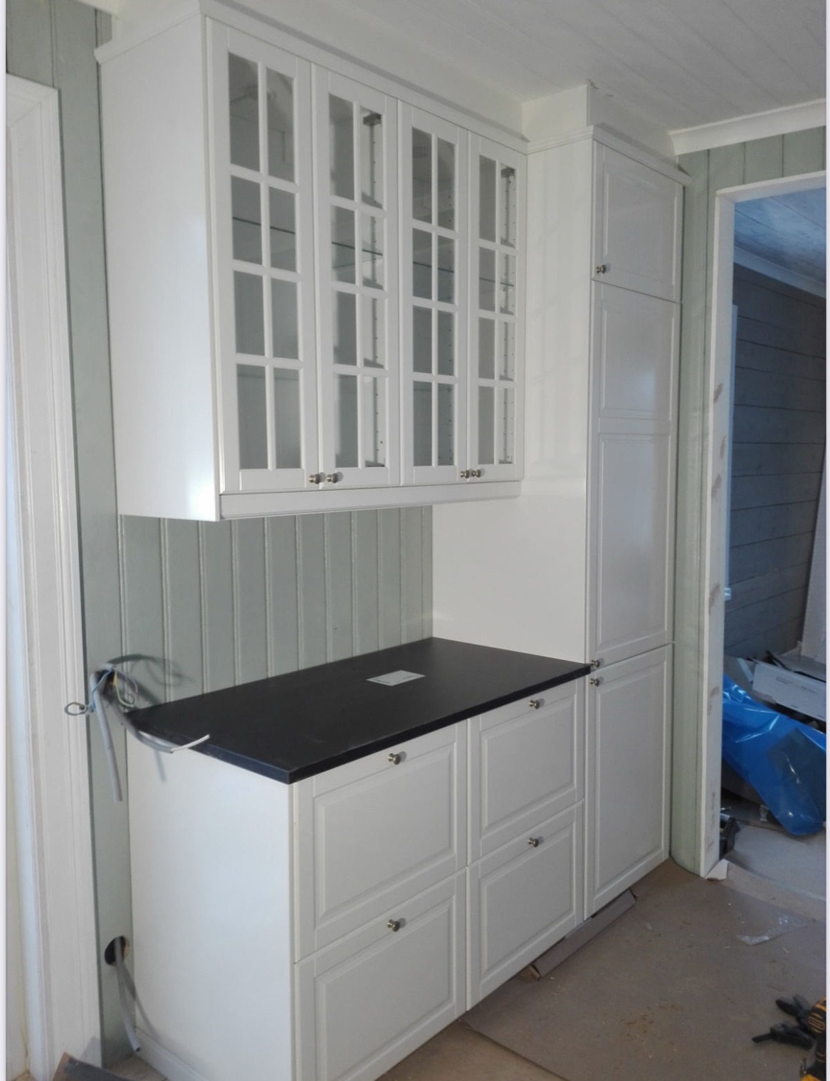 Białe szafki kuchenne z przeszklonymi górnymi drzwiczkami i czarnym blatem, widoczne przewody elektryczne wystające ze ściany.