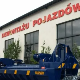 Budynek stacji demontażu pojazd&oacute;w z napisem i niebieską maszyną do transportu wrak&oacute;w na pierwszym planie.