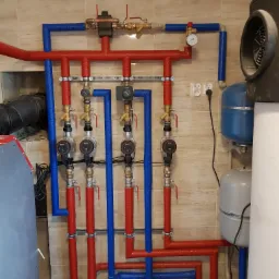 Rozbudowana instalacja kotłowni z kotłem, widoczne czerwone i niebieskie rury z izolacją, pompy obiegowe, zawory, manometr, zbiorniki wyrównawcze i fragment kotła z wentylatorem. Ściana wyłożona...