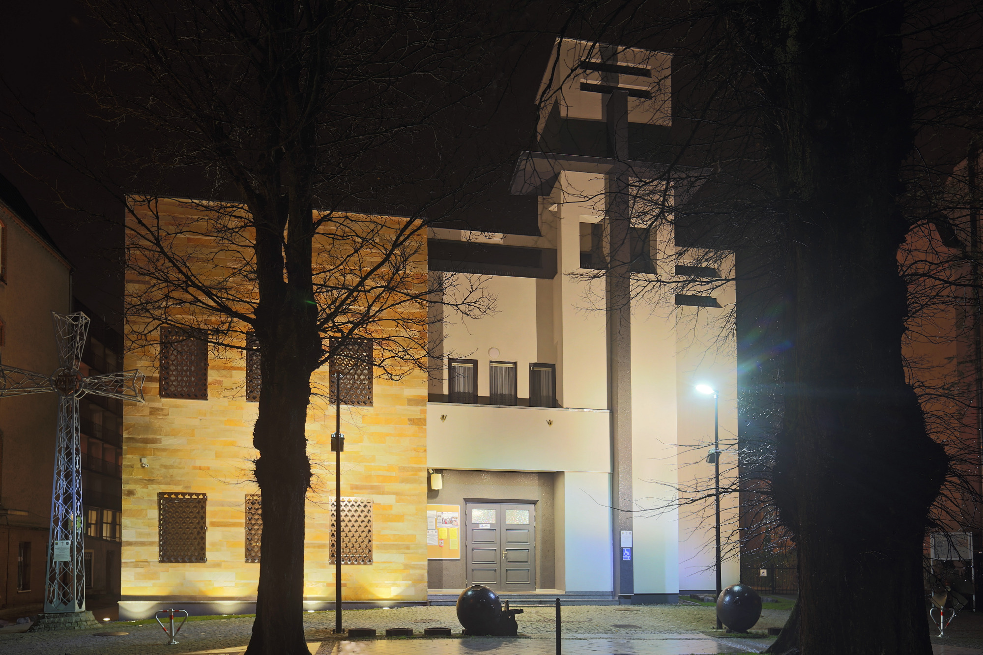 Nocne ujęcie nowoczesnego budynku z elewacją z piaskowca i białymi elementami, oświetlonego latarniami ulicznymi, widoczne drzewa z nagimi gałęziami.