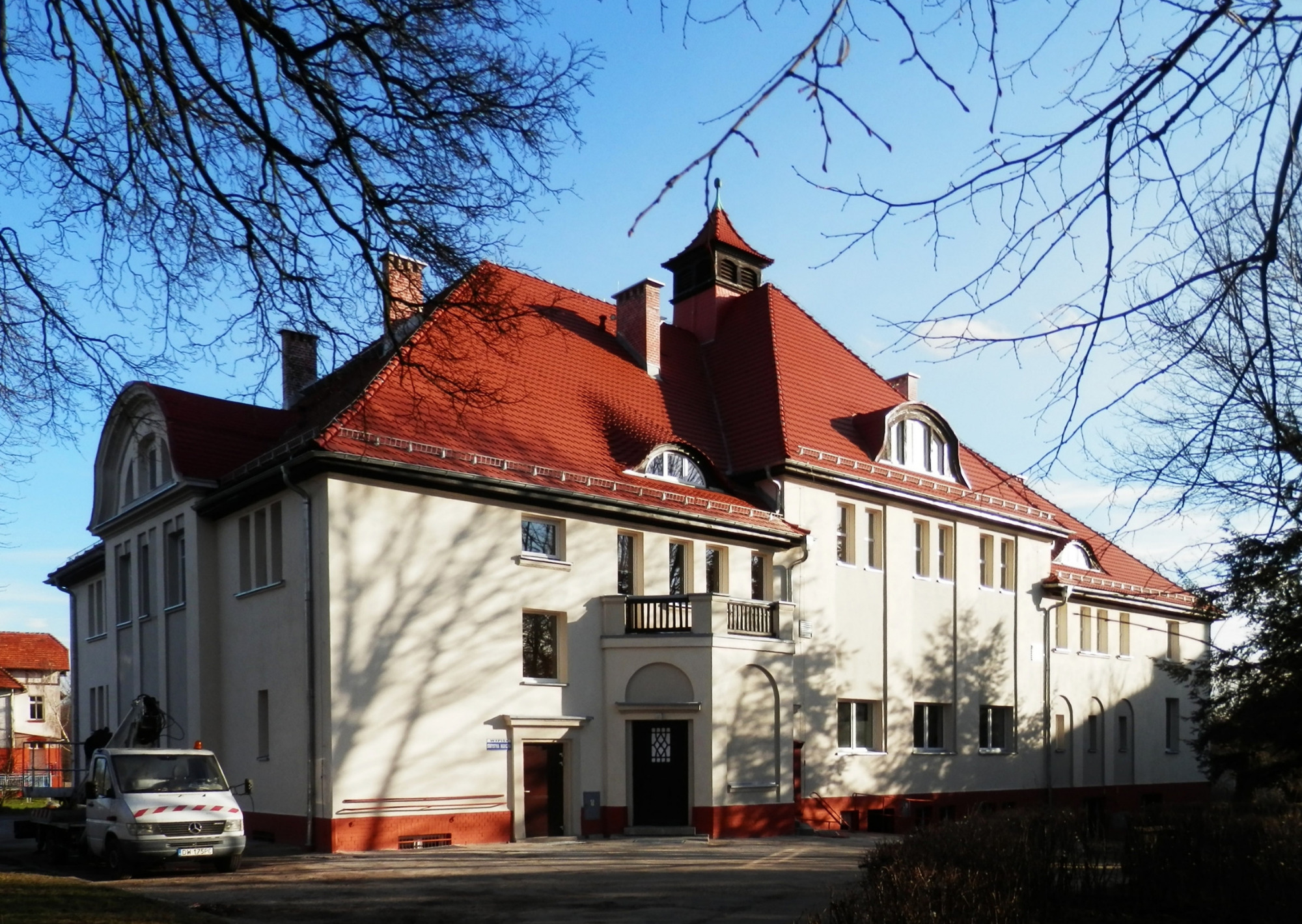 Budynek z czerwoną dachówką w Kłodawie, widoczne ślady cieni drzew na elewacji, z boku zaparkowany biały samochód z podnośnikiem.