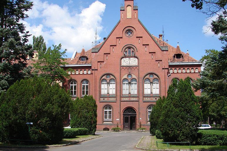 Fasada zabytkowego, ceglanego budynku z czerwonym dachem, widoczna spomiędzy zieleni drzew i krzewów, z elementami dekoracyjnymi i oknami o łukowatych nadprożach.