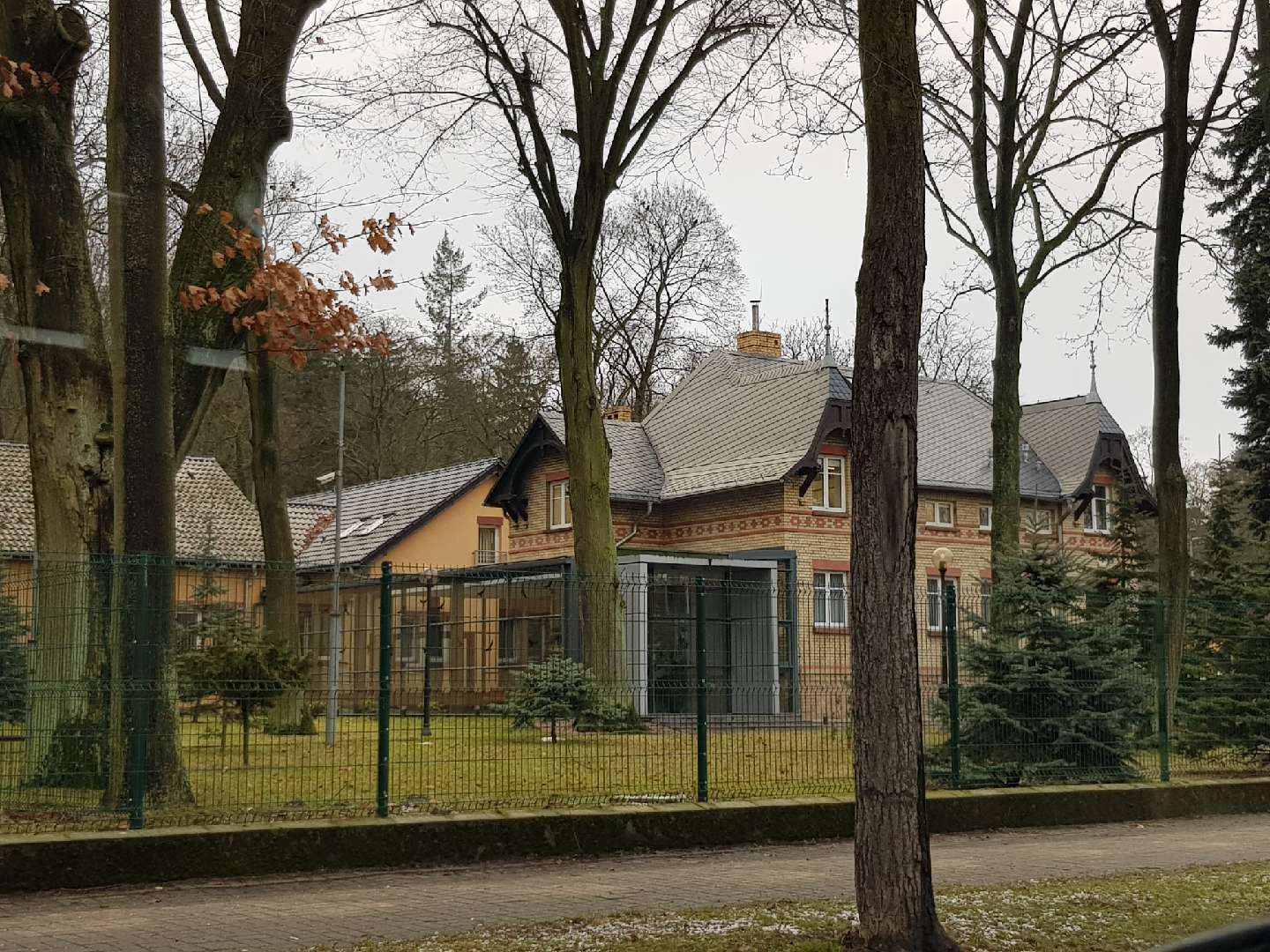 Dwukondygnacyjny budynek mieszkalny z cegły klinkierowej z szarym dachem wielospadowym, widoczny zza drzew i ogrodzenia.