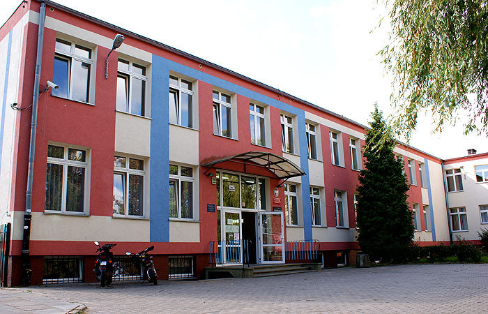 Dwupiętrowy budynek szkoły z czerwono-biało-niebieską elewacją, białymi oknami i przeszklonym wejściem, na pierwszym planie dwa skutery.