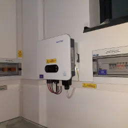 Biały falownik SOFAR zamontowany na ścianie obok dwóch rozdzielnic elektrycznych, widoczne kable i żółte naklejki ostrzegawcze.