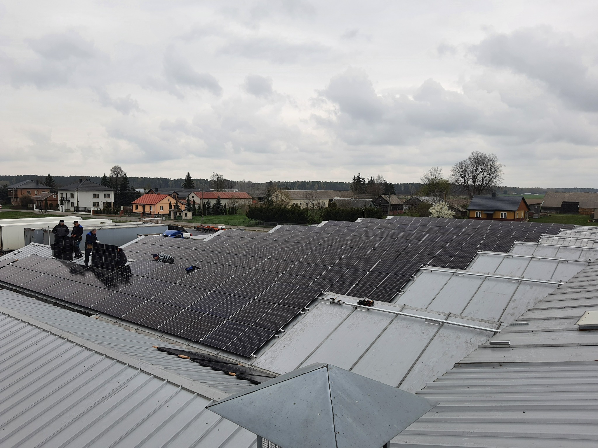 Montaż paneli słonecznych na metalowym dachu budynku gospodarczego, widoczne domy w tle i pochmurne niebo.