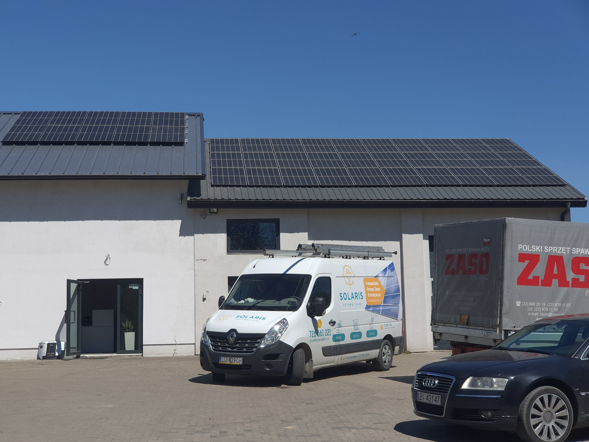 Biały budynek z panelami słonecznymi na dachu, firmowy van Solaris Fotowoltaika z drabiną na dachu zaparkowany przed wejściem, obok zaparkowany czarny samochód osobowy i ciężarówka ZASO.