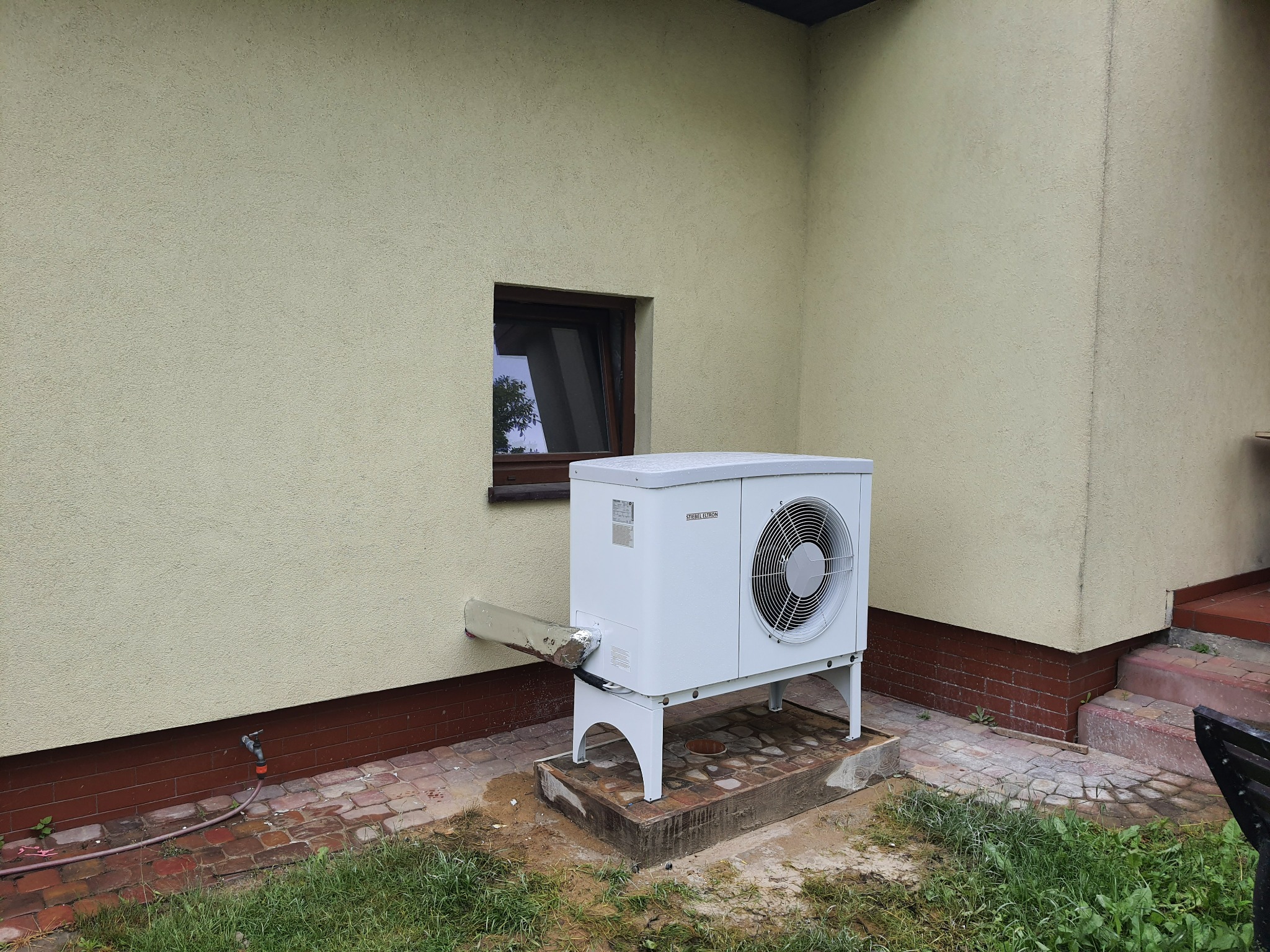 Biała jednostka zewnętrzna pompy ciepła Stiebel Eltron zamontowana przy ścianie budynku z małym oknem, widoczny odpływ skroplin i fragment kostki brukowej.