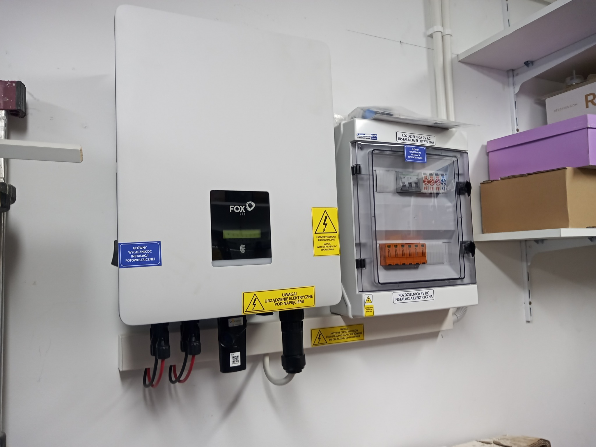 Biały falownik fotowoltaiczny Fox ESS z widocznymi kablami i naklejkami ostrzegawczymi, obok rozdzielnica AC/DC na białej ścianie w pomieszczeniu technicznym z półkami.