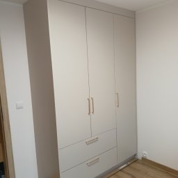 WOPREM - Jasnoszara szafa trzydrzwiowa z szufladami i złotymi uchwytami, dopasowana do wnętrza pokoju z drewnianą podłogą. Widoczny fragment ściany z gniazdkiem elektrycznym.