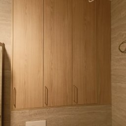 WOPREM - Drewniana szafka łazienkowa z czterema drzwiczkami i złotymi uchwytami, zamontowana nad spłuczką toalety, na tle marmurowej ściany. Widoczne przewody elektryczne.